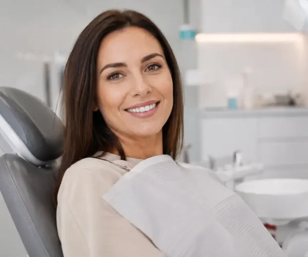 Pacientă adultă zâmbitoare, așezată confortabil pe scaunul stomatologic într-o clinică dentară modernă, luminoasă și elegantă, cu fundal discret blurat și atmosferă calmă și premium.